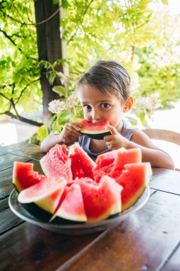 Neşeli bir çocuk, yemyeşil bitkilerle çevrili bir bahçe masasında otururken sulu kırmızı bir karpuzun büyük bir ısırığını alır..