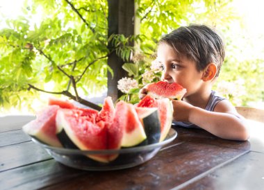 Neşeli bir çocuk, yemyeşil bitkilerle çevrili bir bahçe masasında otururken sulu kırmızı bir karpuzun büyük bir ısırığını alır..
