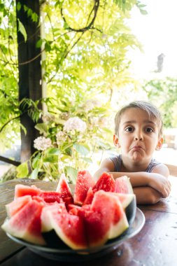 Bahçe masasında oturan bir çocuk, yeşillikle çevrili, içinde komik ya da Çocuk Bakışlı kırmızı bir karpuz dilimi olan Garden Table 'daki SliceS of Watermelon' a isteksizce bakıyor. Sağlıklı Yeme Alışkanlıklarında Gerçek An hoşnutsuz ifade.