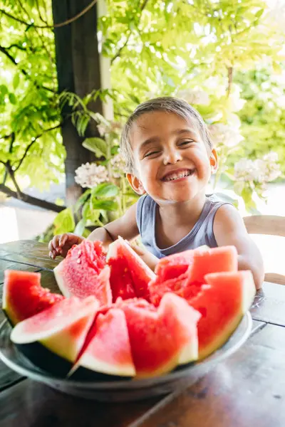 Mutlu bir çocuk, yemyeşil çimlerle çevrili bir bahçe masasında otururken bir dilim parlak kırmızı karpuzun tadını çıkarır..