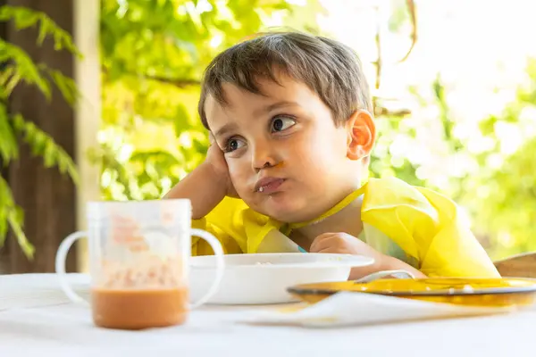 Küçük bir çocuk yemek sırasında masada otururken yorgun ve ilgisiz görünüyor..