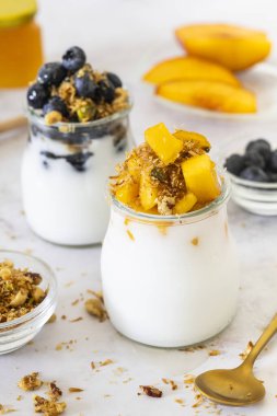 Parlak ve iştah açıcı bir kahvaltı sahnesi iki sağlıklı vegan ve glütensiz seçeneği gösteriyor: biri hindistan cevizli yoğurt, taze şeftali dilimleri ve müsli, diğeri hindistan cevizli yoğurt, yabanmersini ve gevrek müsli.