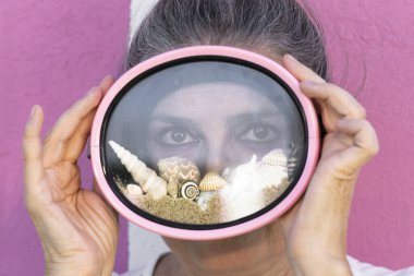 Plaj kumu, deniz kabuğu ve deniz salyangozu dolu pembe dalış gözlüğü takan güzel bir kadının yaratıcı yaz konsept portresi. Yaz tatilinin sembolü, sahil yaşam tarzı, seyahat ve tatil havası.