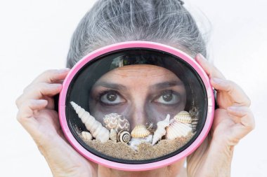 Plaj kumu, deniz kabuğu ve deniz salyangozu dolu pembe dalış gözlüğü takan güzel bir kadının yaratıcı yaz konsept portresi. Yaz tatilinin sembolü, sahil yaşam tarzı, seyahat ve tatil havası.