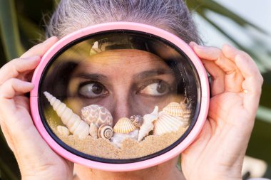 Plaj kumu, deniz kabuğu ve deniz salyangozu dolu pembe dalış gözlüğü takan güzel bir kadının yaratıcı yaz konsept portresi. Yaz tatilinin sembolü, sahil yaşam tarzı, seyahat ve tatil havası.