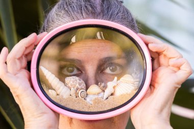 Plaj kumu, deniz kabuğu ve deniz salyangozu dolu pembe dalış gözlüğü takan güzel bir kadının yaratıcı yaz konsept portresi. Yaz tatilinin sembolü, sahil yaşam tarzı, seyahat ve tatil havası.