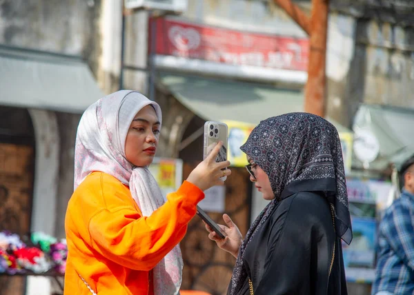 24 Şubat 2023 - George Town - Penang-Malezya - Kafalarında geleneksel bir eşarpla turuncu ve siyah giyinmiş iki genç kadın telefonlarıyla fotoğraf çekiyor..