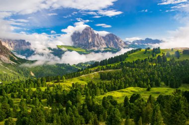 İtalya 'nın Dolomite Alpleri' ndeki Sassolungo Massif ve Gardena Valley 'in canlı bir tasviri. Ön planda yemyeşil bir vadi sergilenirken, arka planda bulutlarla kaplı dağ zirveleri ön plana çıkıyor.
