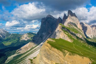 İtalya, Güney Tyrol Dolomiteleri 'ndeki Odle-Geisler Grubu' nun nefes kesici bir görüntüsü. Mavi gökyüzü ve beyaz bulutların arkaplanı resimli kayalarla ve çimlerle kaplı dağ sırasını tamamlıyor.