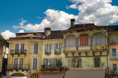 Weathered, colorful facades of traditional Italian buildings in Domodossola's main square, featuring arched windows, balconies, and shutters under a blue sky with fluffy clouds clipart