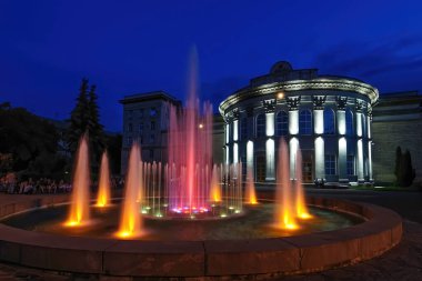Geceleri sütunları olan neoklasik bir binanın önünde renkli ışıklandırılmış çeşme görüntüsü. Sahne, Ukrayna 'nın canlı gece hayatı ve mimarisini yansıtıyor.