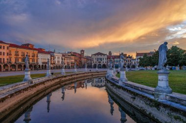 İtalya, Padova 'daki Prato della Valle meydanının güzel günbatımı manzarası, tarihi binalar, heykeller ve renkli bir akşam gökyüzünün altında yansıtıcı bir kanal.