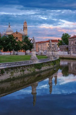 İtalya 'nın Padua kentindeki Prato della Valle Meydanı' nın manzarasında, dramatik bir günbatımı gökyüzü altında Santa Giustina Bazilikası 'nın yansımaları olan bir kanal boyunca heykeller yer alıyor.