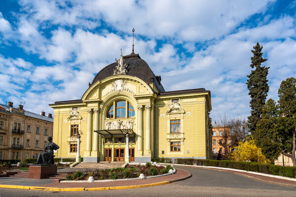 Величественное желтое здание с великолепной архитектурой, с купольной крышей и парадным входом. Статуя стоит на переднем плане, окруженная круговой дорожкой. Черновицкий оперный театр, Украина