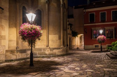 Lviv 'deki Ermeni Katedrali' nin dışındaki sokak lambalarında çiçek sepetleriyle aydınlanmış parke taşı. Tarihsel mimari ve sıcak aydınlatma romantik bir atmosfer yaratır.