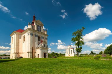 Kırmızı çatılı ve süslü maskeli çarpıcı beyaz Barok kilisesi, kabarık bulutlu canlı mavi gökyüzüne karşı kurulmuş. Tarihsel bina Lopatyn, Ukrayna çevresindeki yemyeşil çimenler ve ağaçlar