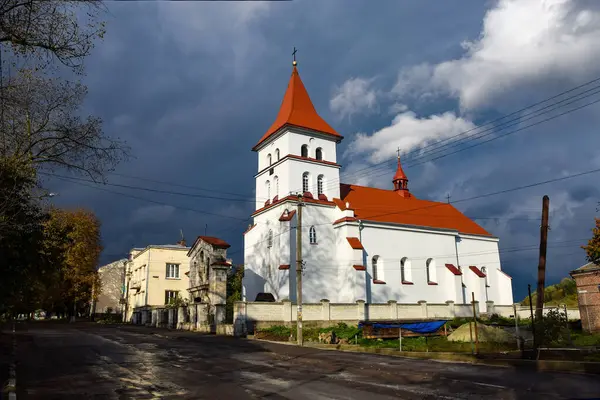 Turuncu çatılı beyaz kilise ve fırtınalı bir gökyüzüne karşı kule. Tarihi bina, çevresindeki binalar ve elektrik hatlarıyla birlikte küçük kasaba ortamında göze çarpıyor. Schyrets, Ukrayna