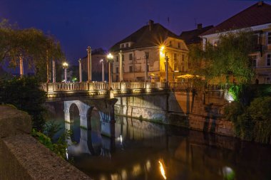 Slovenya 'nın Ljubljana kentinde alacakaranlık sırasında sakin Ljubljanica Nehri' nin üzerinde belirgin sütunlar ve sıcak sokak lambaları bulunan Cobblers Köprüsü,
