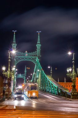 Aydınlatılmış Özgürlük Köprüsü 'nden geçen sarı tramvay Macaristan' ın Budapeşte kentindeki Tuna Nehri üzerinde gece gökyüzüne karşı canlı ışıklar sergiliyor.