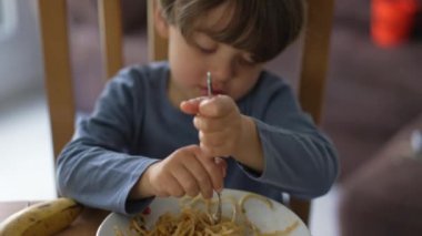 Küçük bir çocuk tek başına çatallı makarna yiyor. Çocuk öğle yemeği yiyor.