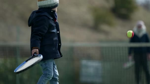 冬の季節に親と一緒にテニスをする子供の背中 コートを着た子供とビーニーがボールを打つ 小さな男の子は屋外でスポーツを — ストック動画