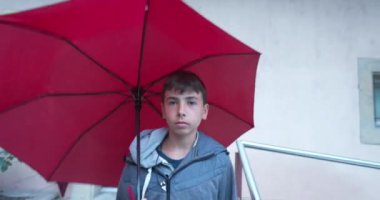 Ergen çocuk elinde şemsiyeyle dışarıda yürüyor. Şehirde öğleden sonra yağmur yağıyor. Genç çocuk yağmurlu bir günde yürür