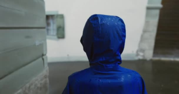 ヨーロッパの街の屋外で雨の中を歩く青いレインコートを着た子供 雨が降りながら歩く子供の背中 — ストック動画