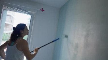 Duvarı uzun çubuklu boya makinesiyle boyayan bir kadın. Kadın evi boyuyor. DIY kavramı