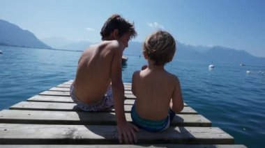 Tahta göl iskelesinde oturan iki küçük çocuk. Büyük kardeş küçük kardeşle takılıyor. Çocuklar dağ kıyısında yaz tatilinin tadını çıkarıyorlar.