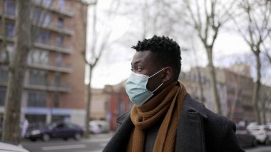 Black African man walking wearing covid surgical face mask in city sidewalk, tracking shot