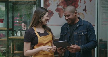 Küçük işletme kahve dükkanının iki farklı çalışanı. Tableti bir arada tutan siyahi bir adam ve önlük giymiş genç bir kadın kameraya gülümsüyor. Kadın ve erkek personel