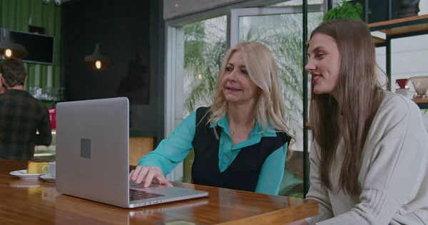 Mature woman entrepreneur speaking with young female staff about work in front of laptop computer. Middle aged senior leader talking about work