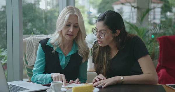 Senior female leader explaining job to younger woman employee at coffee shop. Boss talking about work with colleague showing notes and computer screen working remotely at cafe restaurant