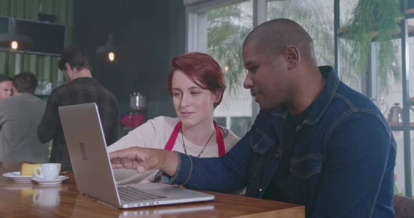 African American manager explaining job to new young female employee in front of laptop sitting at coffee shop. Two diverse staff collaborating together using computer. A black man talking with woman co worker