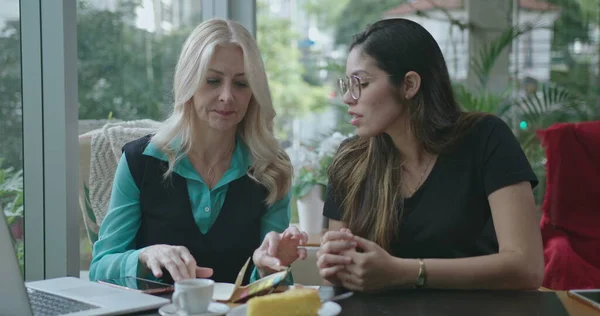 Senior female leader explaining job to younger woman employee at coffee shop. Boss talking about work with colleague showing notes and computer screen working remotely at cafe restaurant