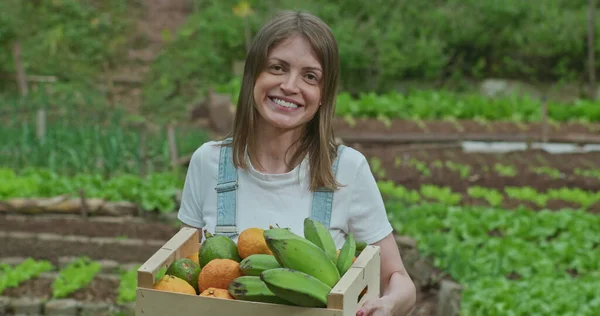 Young happy woman holding basket of fruits and vegetables outside in green field. A female farmer owner of small business holds bananas and tangerines