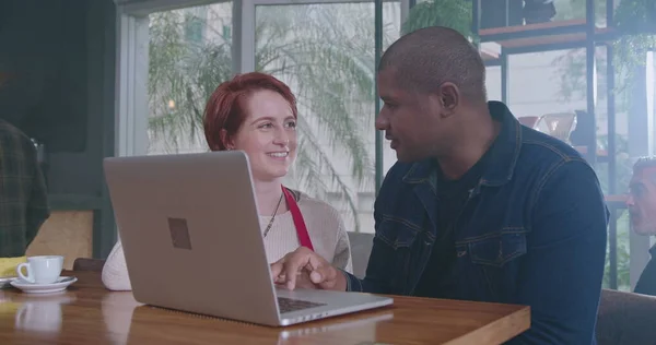 African American manager explaining job to new young female employee in front of laptop sitting at coffee shop. Two diverse staff collaborating together using computer. A black man talking with woman co worker