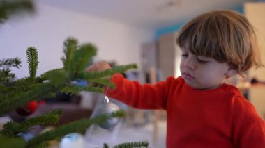 Anne çocuğa Noel süslerini ağaca asması için yardım ediyor. Kış mevsimi şenlikleri. Yıllık gelenek