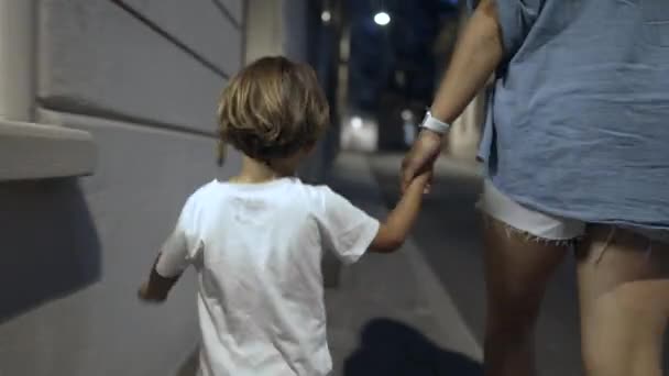 夜の歩道で街を歩く母親と子供の手を握っている 小さな男の子の後ろに親と夕方に屋外に歩く — ストック動画