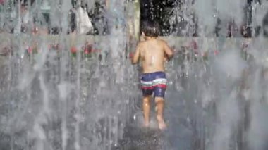 Küçük çocuk yaz günü şehir parkında su jetlerinde koşuyor. Küçük çocuk ferahlatıcı suda koşar yavaş çekimde 120 fp sıçrar.