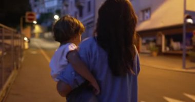 Anne, geceleri kaldırımda yürürken kucağında çocuk taşıyor. Ebeveynin sırtında kucağında küçük bir çocuk akşam yürür.