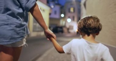 Anne ve çocuk el ele tutuşuyorlar. Gece şehir caddesinde yürüyorlar. Anne ve çocuk el ele tutuşup kaldırımda yürüyorlar.