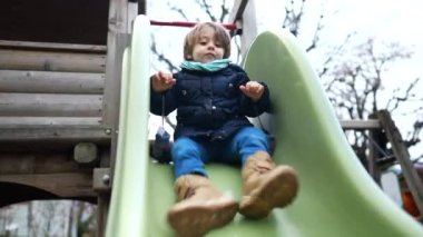 Çocuk kış mevsiminde oyun parkında kayıyor. Palto ve eşarp giyen küçük bir çocuk dışarıda eğleniyor.