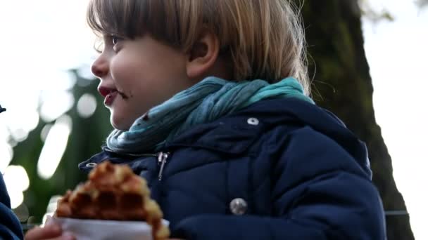 一个快乐的小男孩在外面吃比利时华夫饼干 可爱的男孩一边吃街头食物一边笑着 — 图库视频影像