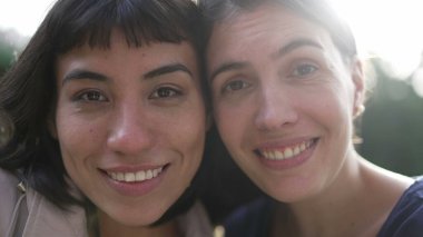 Two young women faces together cheek to cheek. Portrait of diverse people looking at camera with sunlight