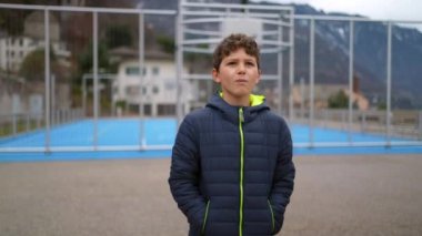 Düşünceli genç bir çocuk şehir parkının dışında, basketbol sahasının yanında elleri ceketinin cebinde duruyor. Düşünceli ifade