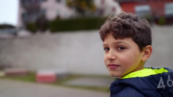 小男孩站在室外看着摄像机在跟踪拍摄 穿着冬季夹克站在公园里的十几岁以下男孩的画像 — 图库视频影像