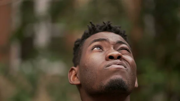 Pensive black African man standing outdoors thinking, looking up at sky in contemplation