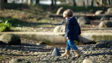 Şehir parkında oynayan kaygısız küçük bir çocuk. Kışlık giysiler ve botlarla doğayı keşfeden bir çocuk. Çocuk taşların üzerinde zıplıyor ve duruyor