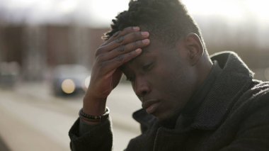 Kafası meşgul siyah Afrikalı adam şehir kaldırımında oturuyor. Endişe duyuyor.